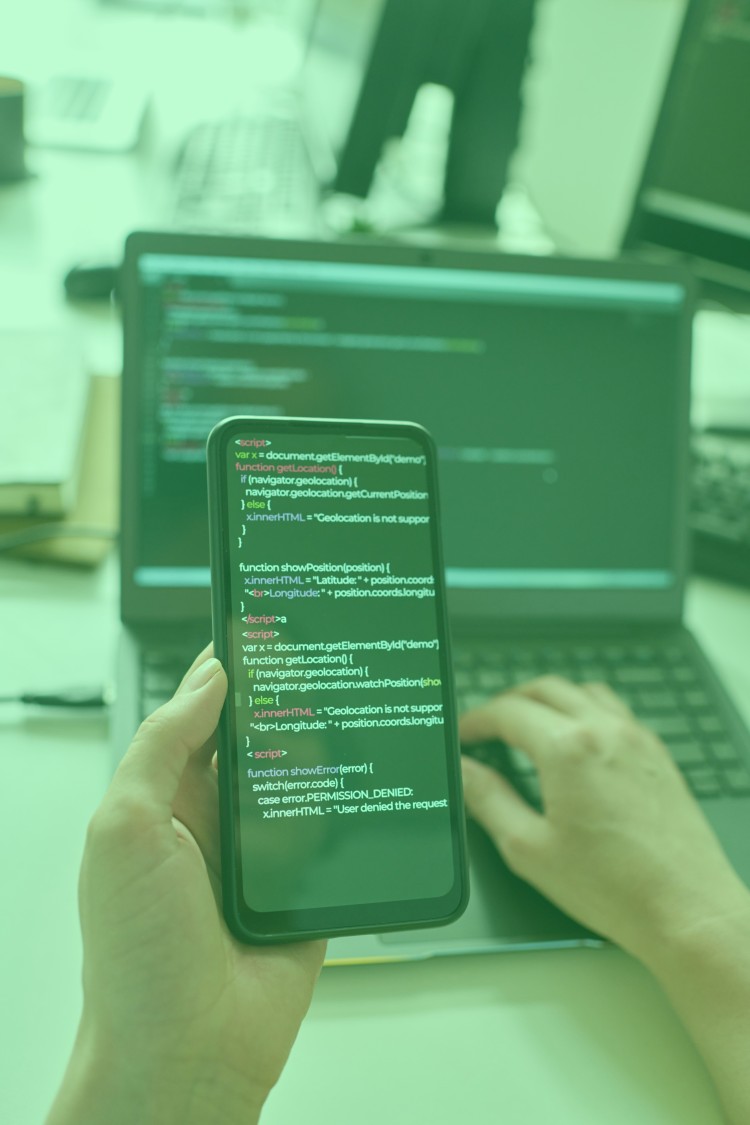 A person holding a smartphone displaying code, with a laptop showing similar code in the background.